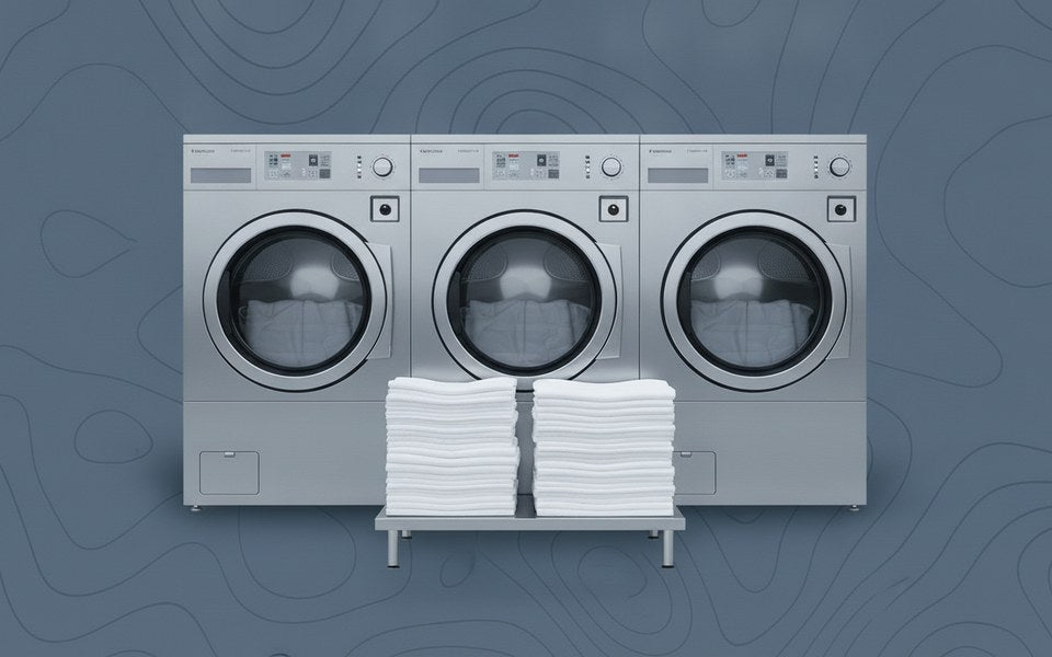 Row of large stainless steel industrial front-load washing machines behind stacks of bright white folded linens