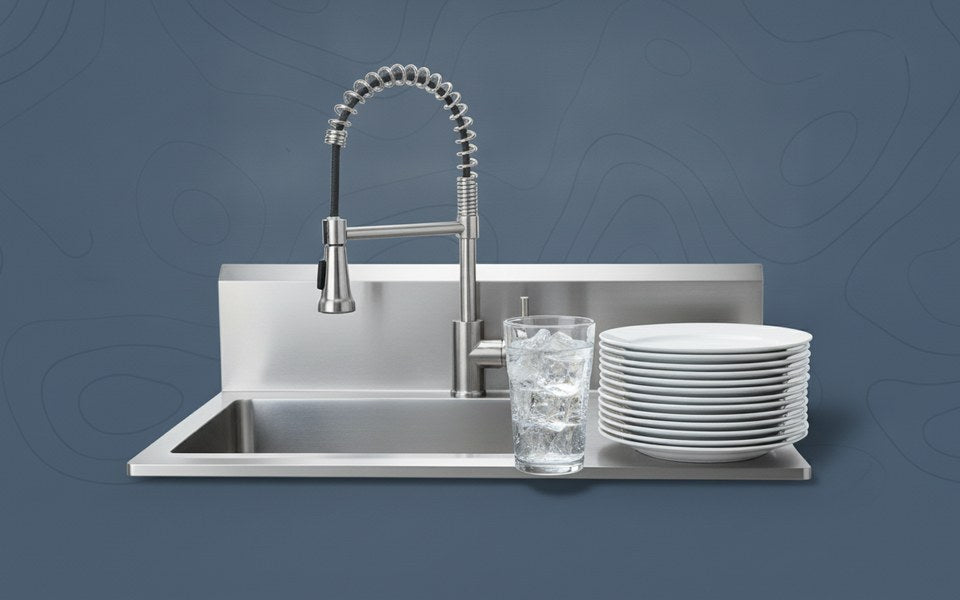 Stainless steel commercial kitchen sink with a pre-rinse spray faucet, a glass of clear iced water and a stack of spotless plates
